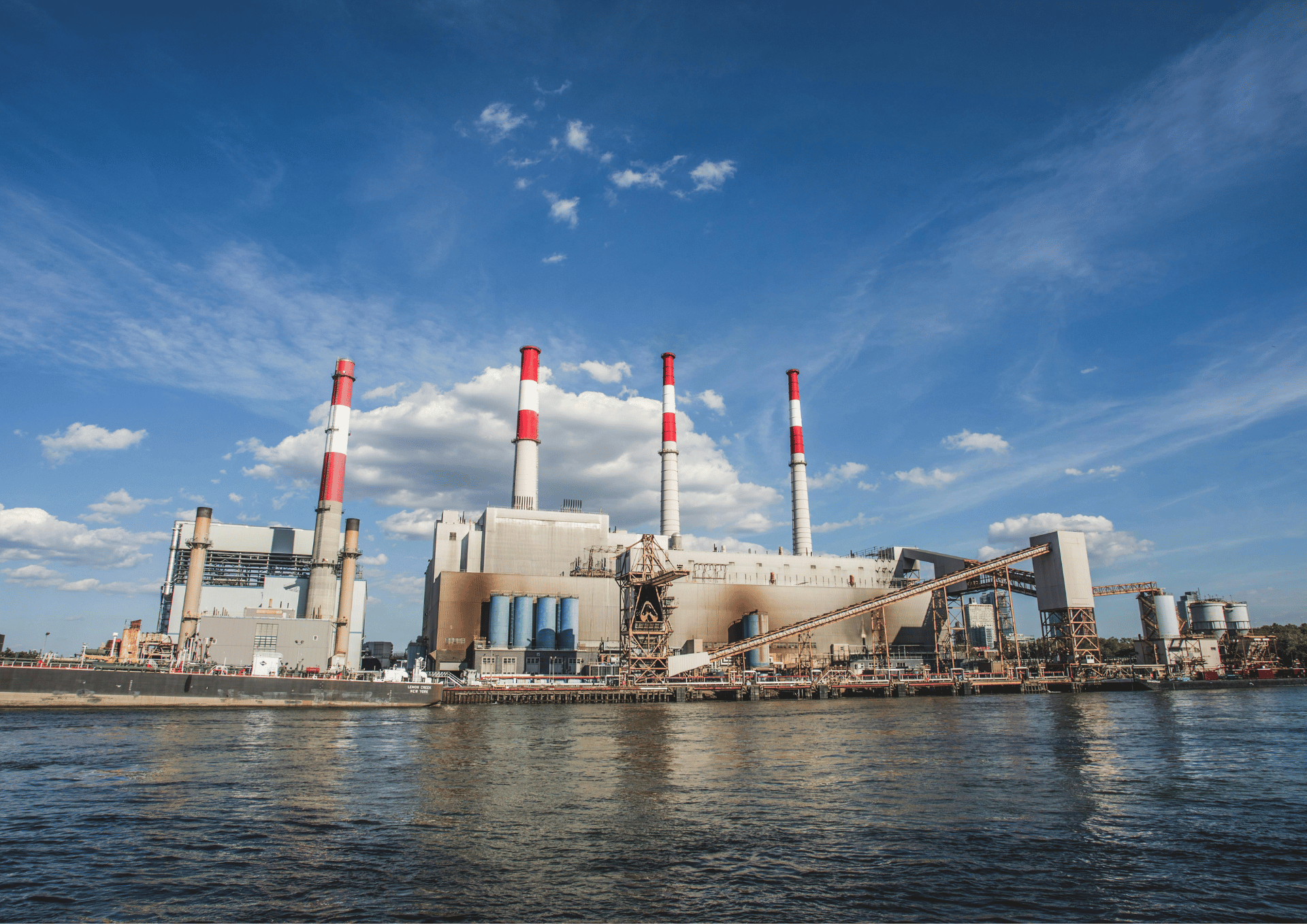 Industrial power plant along a river with tall red-and-white chimneys under a blue sky.