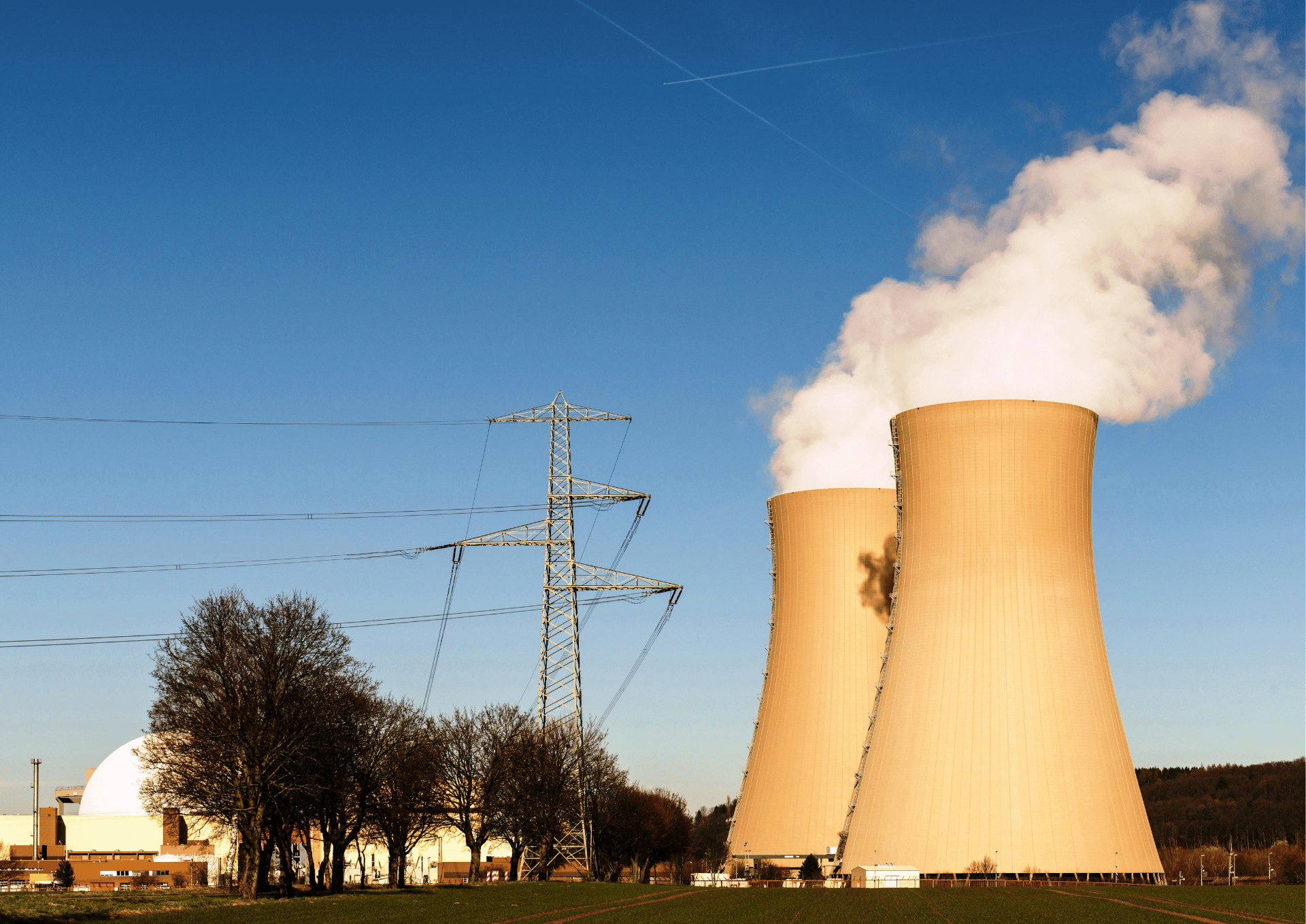 Two beige cooling towers emit steam as power lines and a transmission tower stand in the foreground against a clear blue sky.