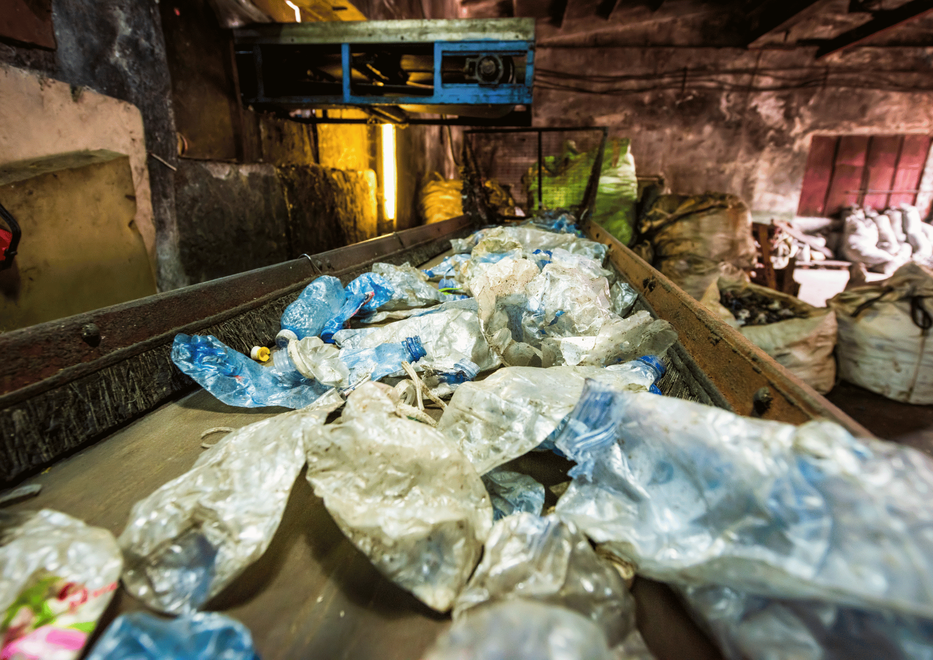 Conveyor belt in a waste facility carrying crumpled plastic bags and litter in a dim, industrial workspace.