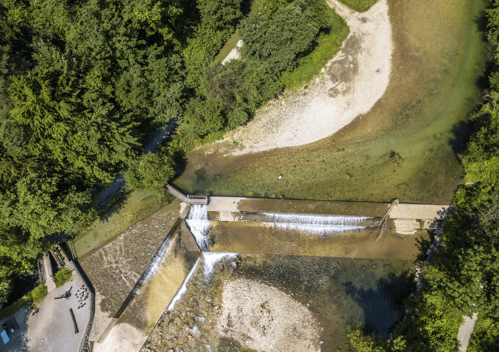 Aerial view of a dam with water flowing over stepped barriers, surrounded by rocky shore and dense green trees on a river.