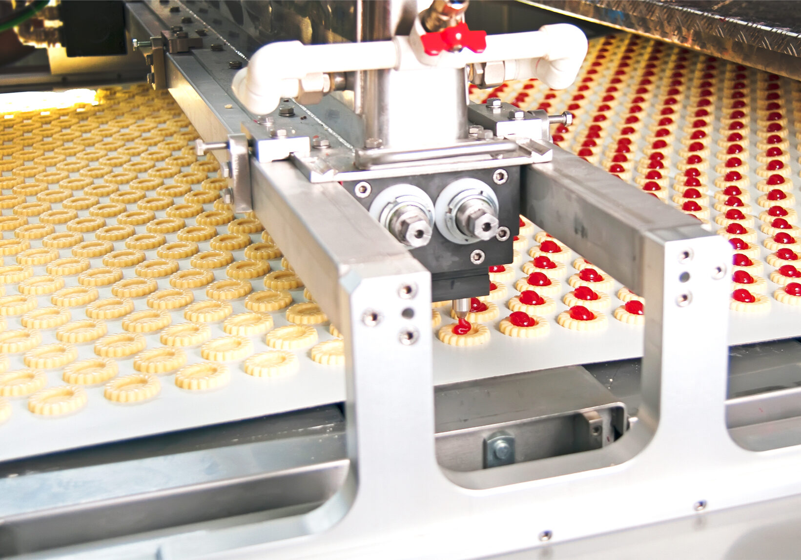 Automated bakery line with ring-shaped cookies moving along a conveyor under metal machinery; some have red centers.